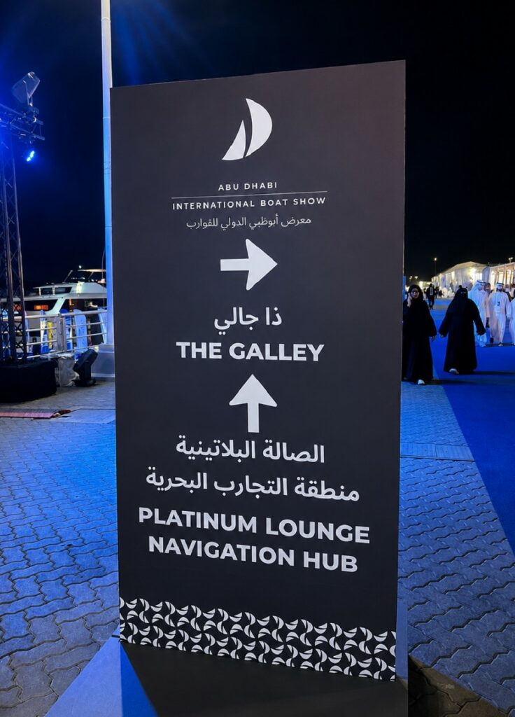 Tótem de señalización nocturna del evento con textos en árabe e inglés ("The Galley", "Platinum Lounge", "Navigation Hub") y el logotipo de vela