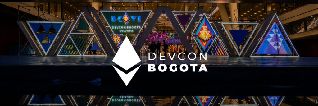 Fotografía de señalización triangular iluminada con el diamante de Ethereum en un recinto, con el logotipo "Devcon Bogotá" superpuesto