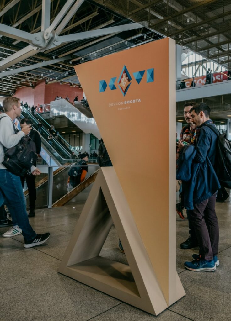 Tótem de señalización triangular en el recinto del evento, con personas alrededor