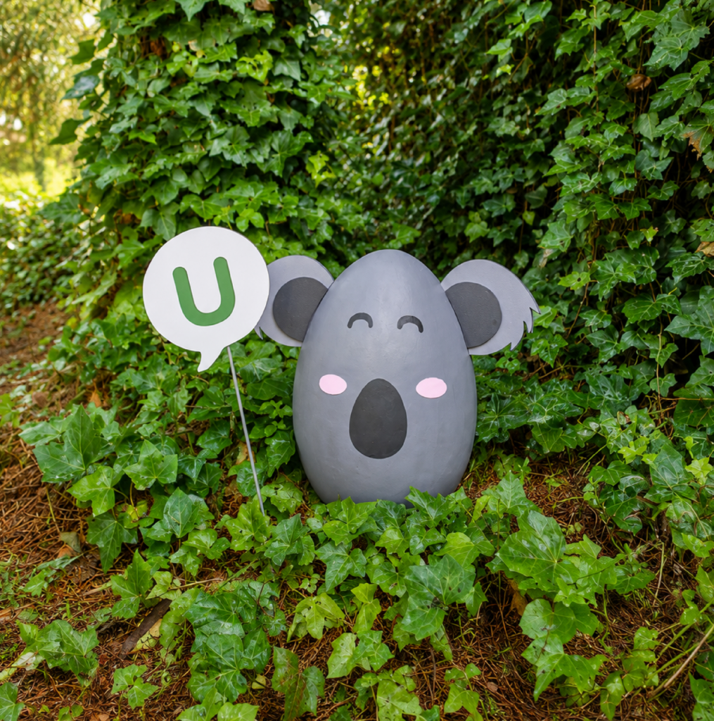 Huevo escultórico con forma de koala escondido entre setos verdes, con un cartel con la letra U