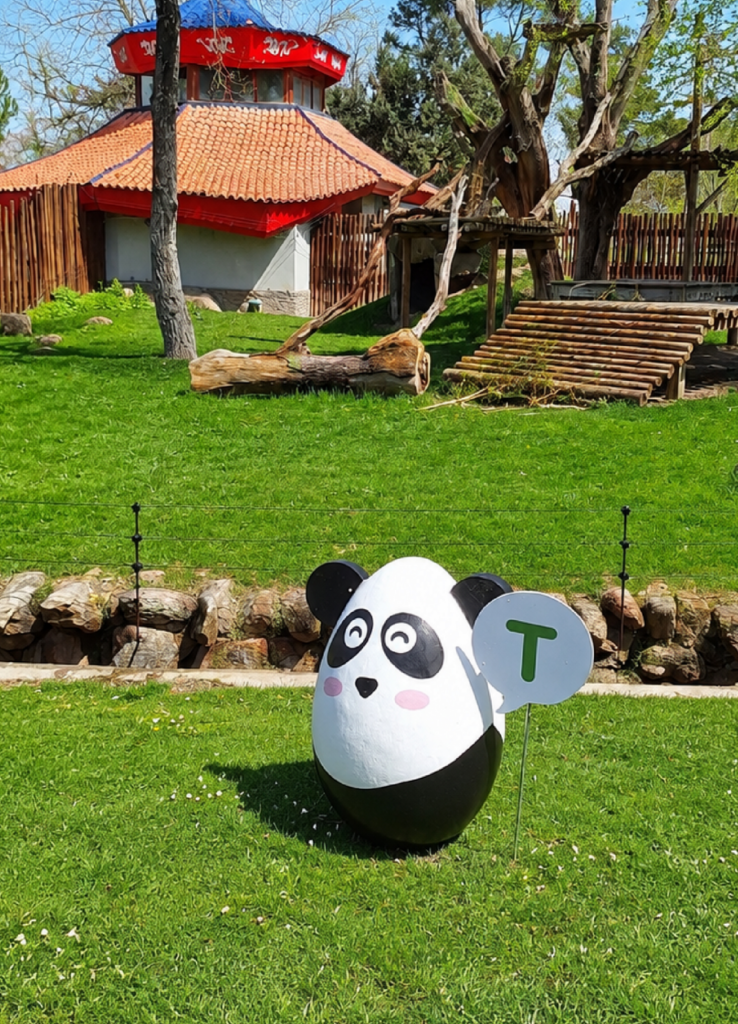 Huevo escultórico con forma de panda sobre el césped junto al pabellón del panda, con un cartel con la letra T