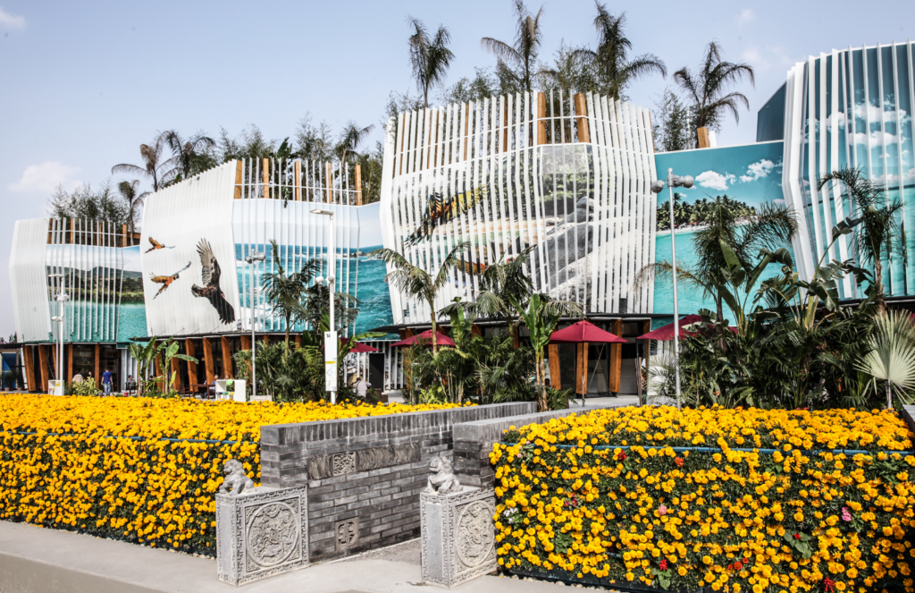 Fachada exterior del pabellón de Colombia con murales de fauna marina y aves entre paneles verticales blancos, plantas tropicales y parterres de flores amarillas
