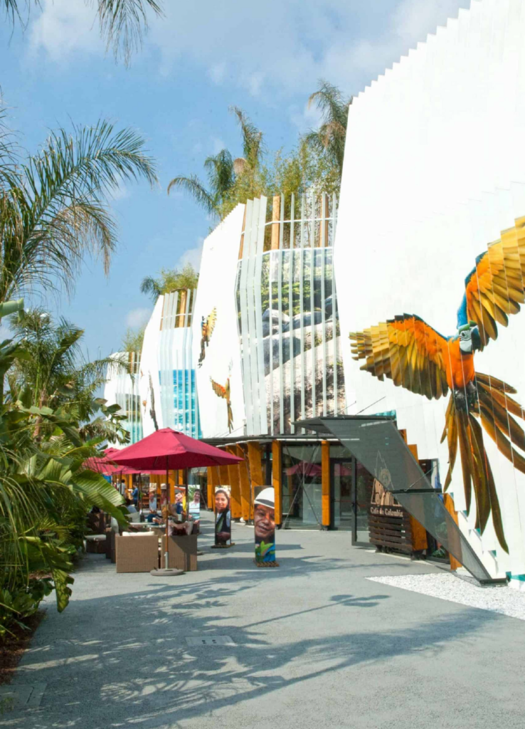 Vista a pie de calle del pabellón con visitantes, una zona de cafetería con sombrilla roja, palmeras y un guacamayo pintado en la fachada