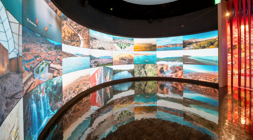Pared curva de pantallas con un mosaico de fotografías aéreas de paisajes peruanos —ciudades, costas y montañas— reflejadas en el suelo