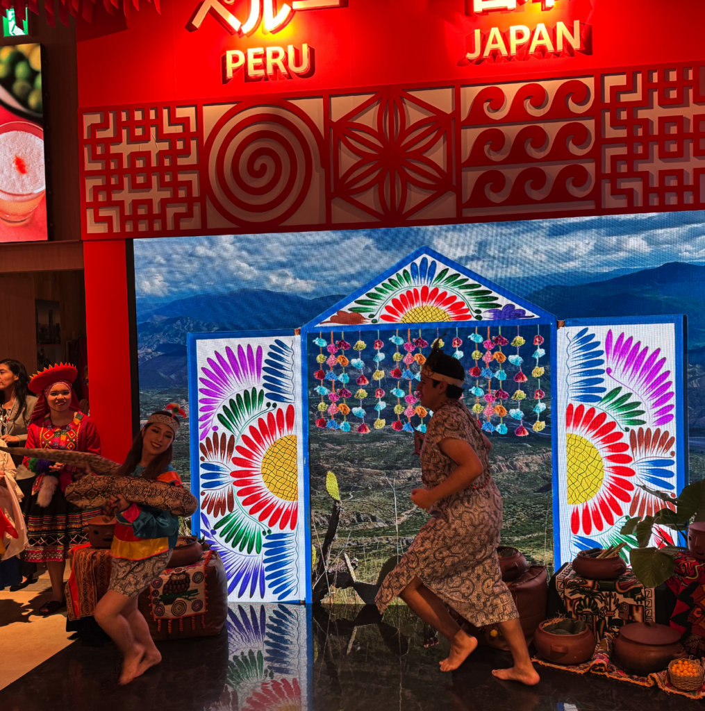 Entrada del pabellón decorada con motivos textiles andinos de flores, con bailarinas en traje tradicional bajo el cartel "Peru Japan"