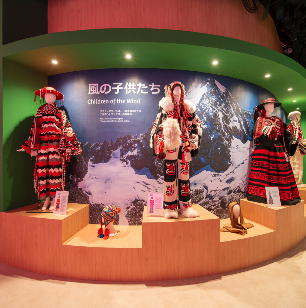 Exhibición "Children of the Wind" con trajes textiles andinos tradicionales sobre maniquíes, con un fondo de montañas nevadas
